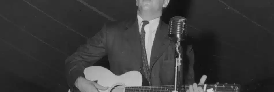 Alan Lomax performing with an acoustic guitar beside a vintage microphone at the Mountain Music Festival around 1938