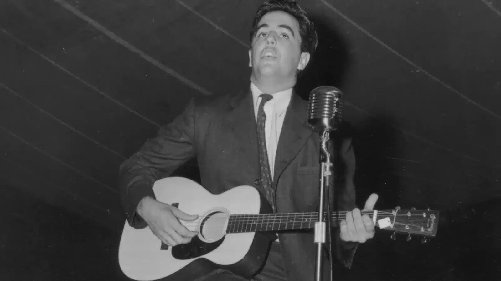 Alan Lomax performing with an acoustic guitar beside a vintage microphone at the Mountain Music Festival around 1938